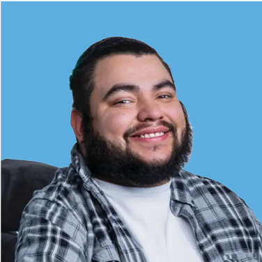 Smiling person with a beard and slicked-back hair, wearing a plaid shirt and white turtleneck, against a light blue background.