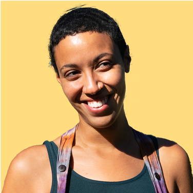 Smiling person with short curly hair, wearing a colorful strap over their shoulders, against a light yellow background.