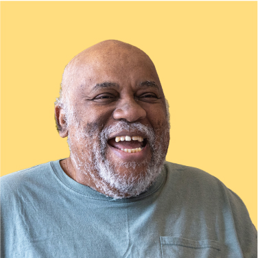 Smiling person with a bald head and a beard, wearing a grey shirt, against a light yellow background.